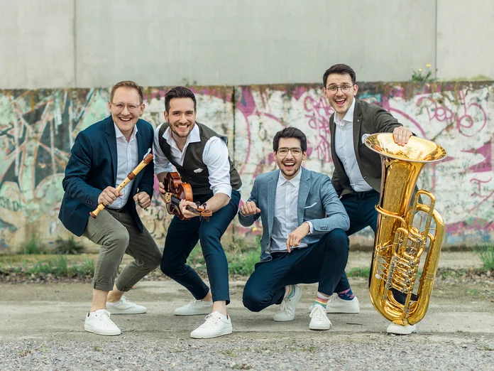 Vier fröhliche Männer (Hanke Brothers) posieren mit Musikinstrumenten vor einer Wand mit Graffiti. Sie wirken energiegeladen und humorvoll.