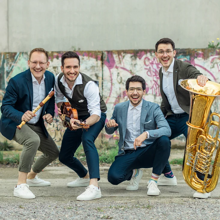 Vier fröhliche Männer (Hanke Brothers) posieren mit Musikinstrumenten vor einer Wand mit Graffiti. Sie wirken energiegeladen und humorvoll.