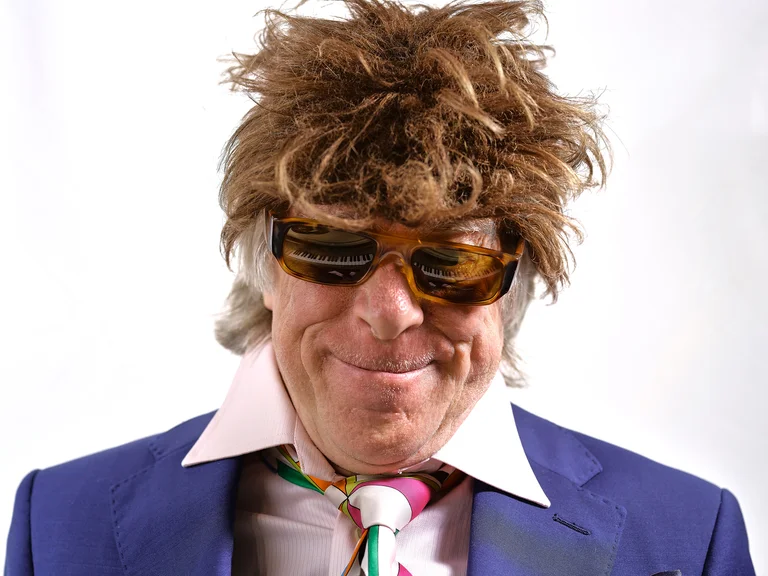 Ein Mann trägt eine auffällige Perücke, eine Sonnenbrille und einen blauen Anzug mit gemusterter Krawatte. Er lächelt.