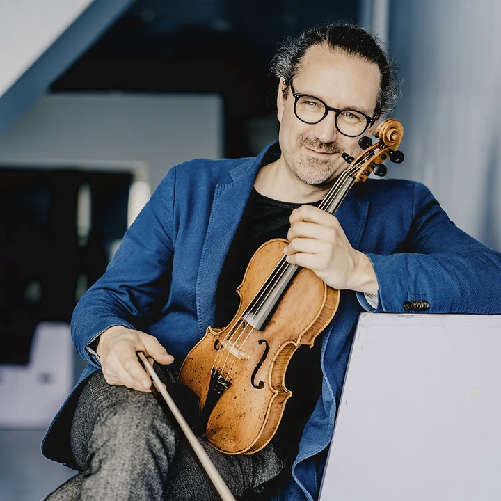 Julien Chauvin sits on a modern piece of furniture with his legs crossed, wearing a blue blazer. He holds his violin and looks into the camera with a smile.