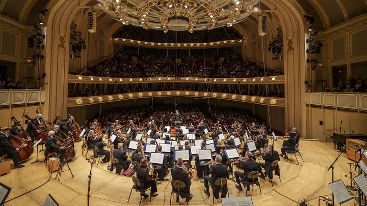 Ein Orchester (Klaus Mäkelä mit dem Chicago Symphony Orchestra) spielt in einem großen, eleganten Konzertsaal mit prachtvoller Beleuchtung und einem Publikum auf mehreren Ebenen.