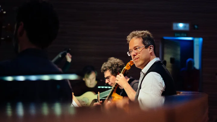 Ein Musiker (Leonardo García-Alarcón) spielt ein Streichinstrument in einem Konzertsaal. Andere Musiker sind im Hintergrund mit Saiteninstrumenten zu sehen.