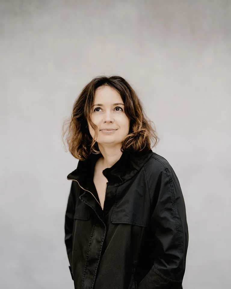 Frau (Patricia Kopatchinskaja) in schwarzem Mantel vor neutralem Hintergrund, die nachdenklich in die Ferne blickt. Leicht welliges Haar und entspannte Haltung.