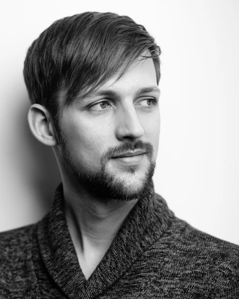 Ein Mann (Felix Schirmer) mit kurzem Haar und Bart schaut nach rechts. Er trägt einen grob gestrickten Pullover. Schwarz-Weiß-Fotografie.