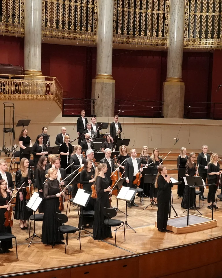 Ein Orchester spielt auf einer großen Bühne in einem eleganten Konzertsaal mit hohen Säulen. Dirigent steht vor den Musikern.