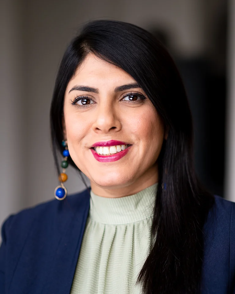 Frau mit dunklen Haaren, blauem Blazer und grünem Oberteil lächelt in die Kamera. Sie trägt markante Ohrringe.