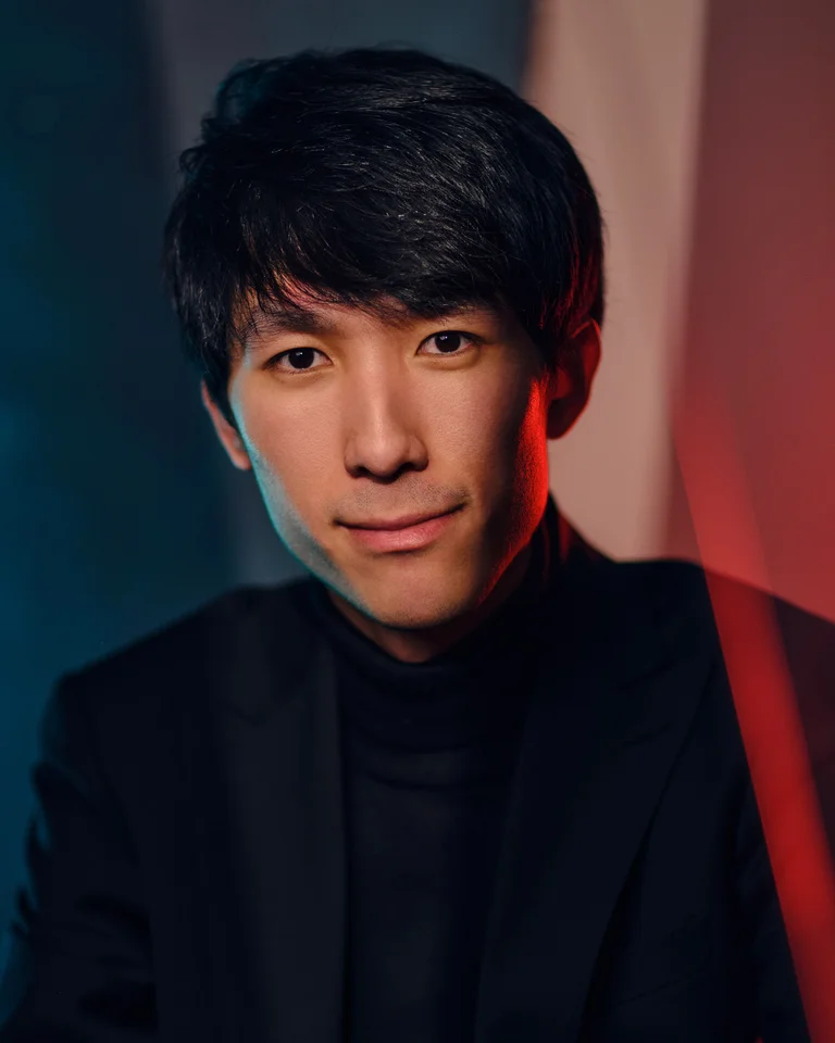 Porträt eines Mannes (Eric Lu) in einem dunklen Anzug vor einem gemusterten Hintergrund mit roter und blauer Beleuchtung.