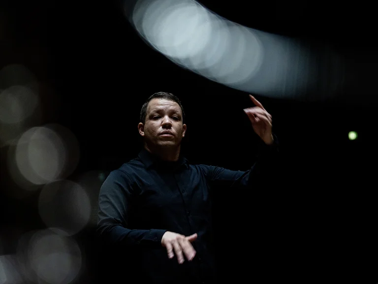 Ein Dirigent (Ryan Bancroft) in einem schwarzen Outfit dirigiert mit ausdrucksvollen Gesten vor einem dunklen Hintergrund mit verschwommenen Lichtern.