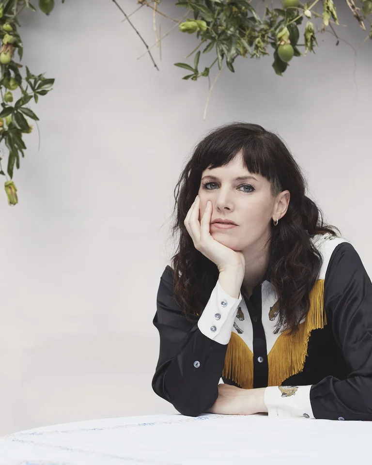Anna Meredith sits at a table, surrounded by plants, resting her chin on her forearm.