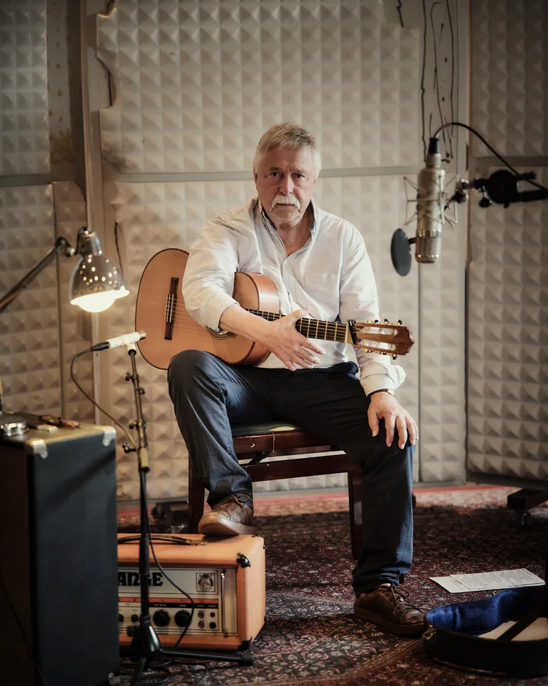 Ein Mann (Wolf Biermann) sitzt in einem Tonstudio mit einer Akustikgitarre. Umgeben von Mikrofonen und Verstärkern, trägt er ein weißes Hemd.