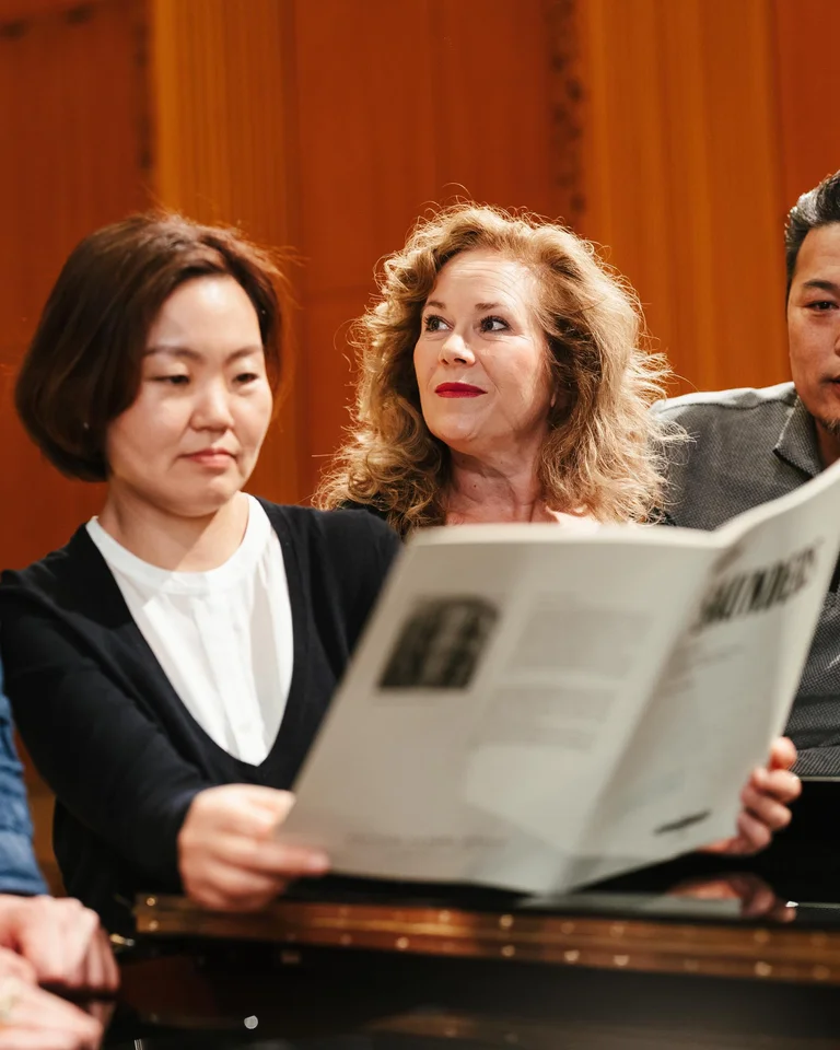 Four people sit together at a piano, looking at sheet music, seemingly planning a musical rehearsal.