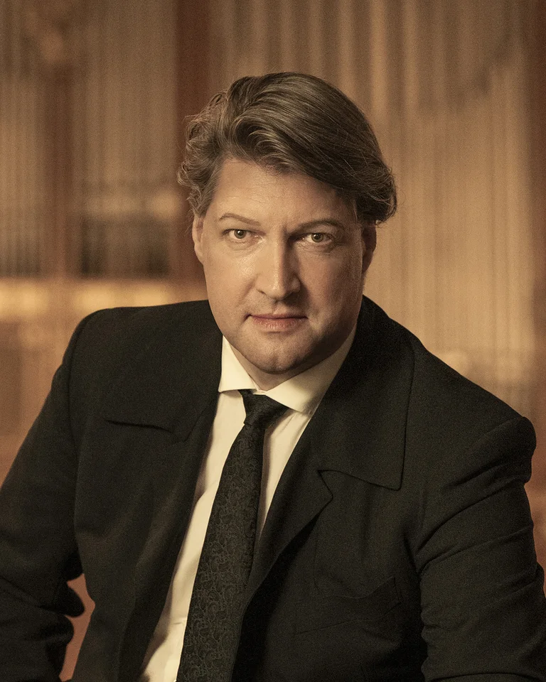 A man (Gunther Rost) in a black suit stands in front of a blurred background resembling a concert hall, with golden light accents.