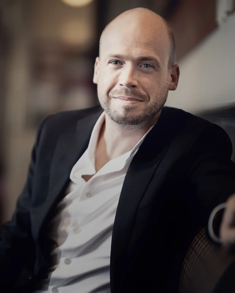 Ein lächelnder Mann (Maarten Engeltjes) in einem dunklen Anzug sitzt entspannt in einem weich beleuchteten Raum, der elegant und einladend wirkt.