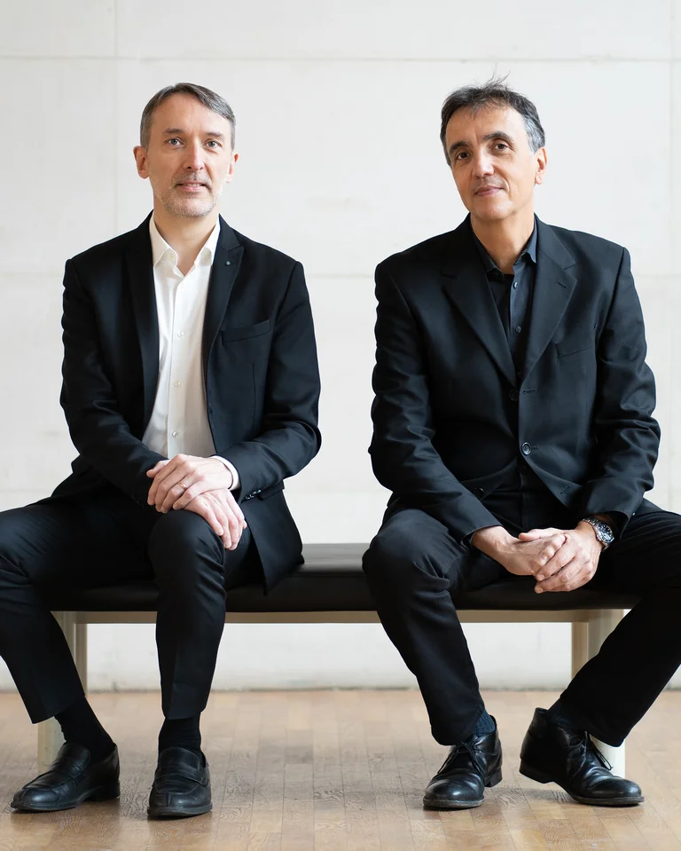 Zwei Männer in eleganten, dunklen Anzügen sitzen nebeneinander auf einer Bank vor neutralem Hintergrund, in einem minimalistischen Raum.