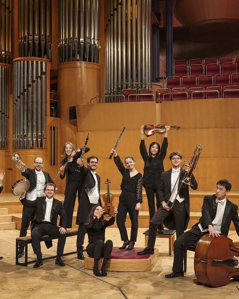Ein Orchester posiert auf einer Bühne mit Holzinstrumenten und formeller Kleidung in einem Konzertsaal der Kölner Philharmonie.