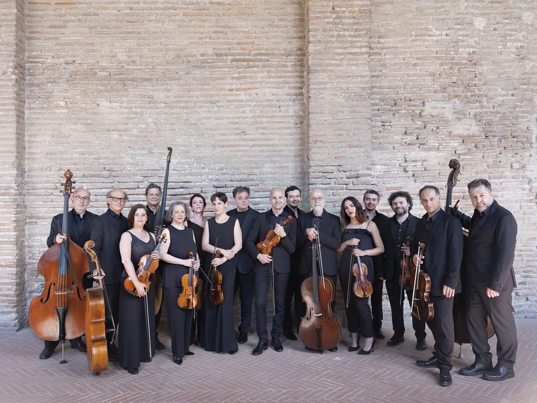 Eine Gruppe von Musikern (Accademia Bizantina) in schwarzer Kleidung posiert mit Streichinstrumenten vor einer Backsteinwand. Sie wirken professionell.