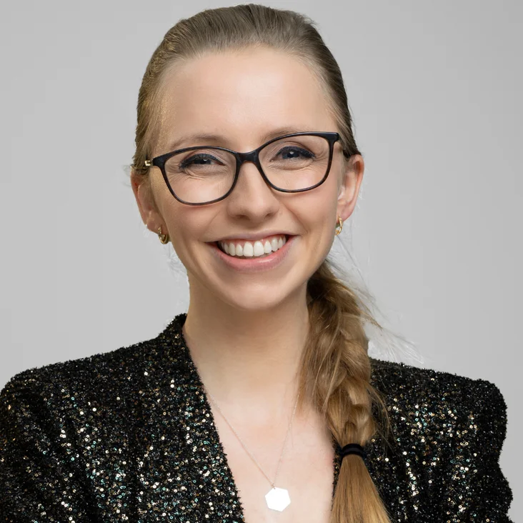 Eine lächelnde Person mit Brille und Zopf trägt einen glitzernden schwarzen Blazer vor einem grauen Hintergrund.