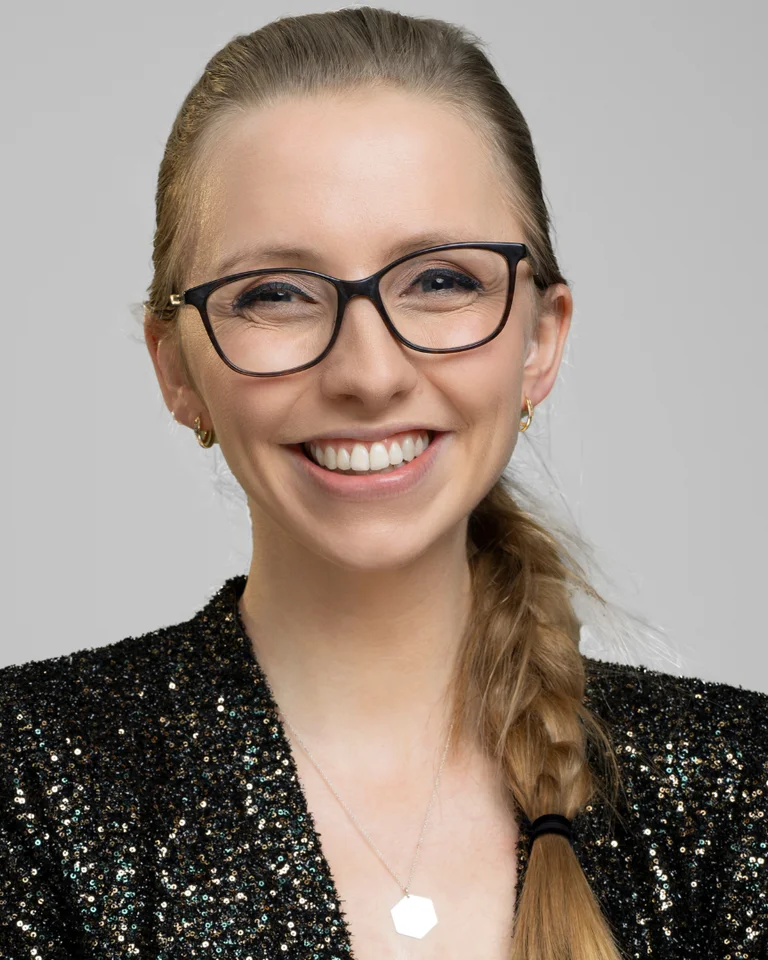 Eine lächelnde Person mit Brille und Zopf trägt einen glitzernden schwarzen Blazer vor einem grauen Hintergrund.