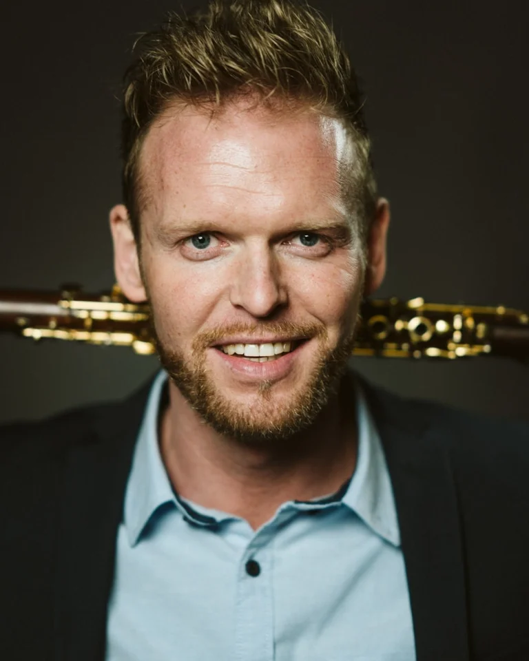 A bearded man in a suit holds a woodwind instrument resting on his shoulders, smiling at the camera.