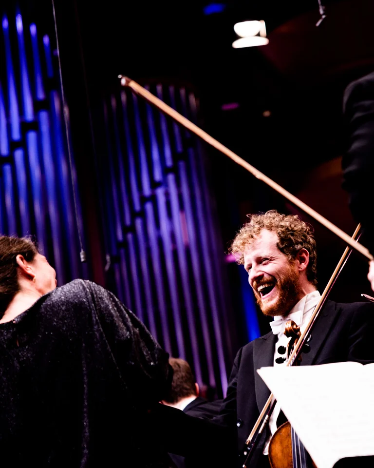 Ein lächelnder Violinist auf der Bühne, umgeben von anderen Musikern. Die Atmosphäre ist warm, mit intensiver Beleuchtung auf vertikalen Linien.