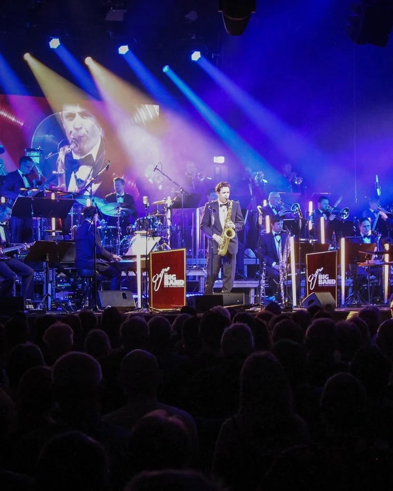 Eine Bühne voller Musiker (Big Band der Bundeswehr) mit verschiedenen Instrumenten, beleuchtet in bunten Lichtern, während ein Konzert mit großem Publikum stattfindet.
