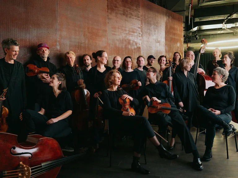 Ein Ensemble von Musikern (Ensemble Resonanz) sitzt mit ihren Streichinstrumenten in einem Proberaum. Sie tragen schwarze Kleidung und wirken konzentriert.