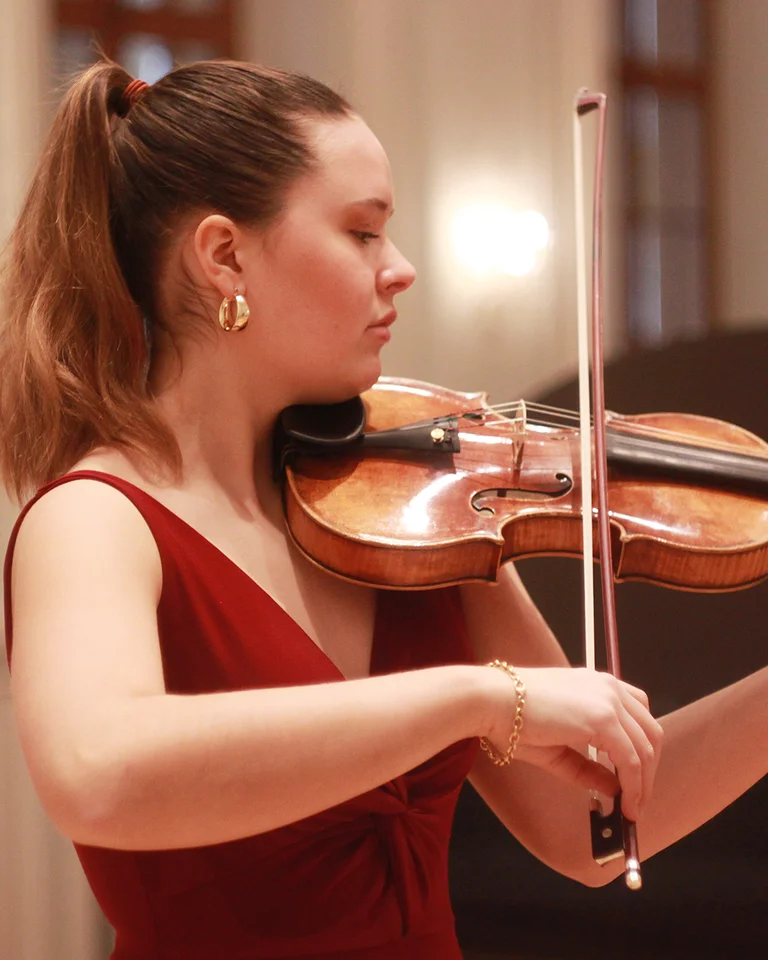 Eine Frau in einem roten Kleid spielt konzentriert Geige in einem eleganten Konzertsaal mit klassischen weißen Wänden.