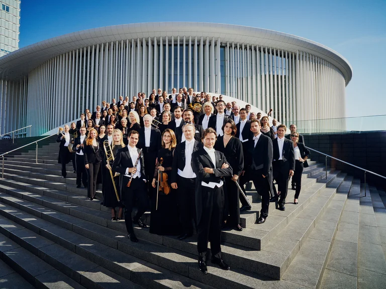 Eine Orchester (Luxembourg Philharmonic) in formeller Kleidung steht auf einer breiten Treppe vor einem modernen, runden Gebäudeeingang.