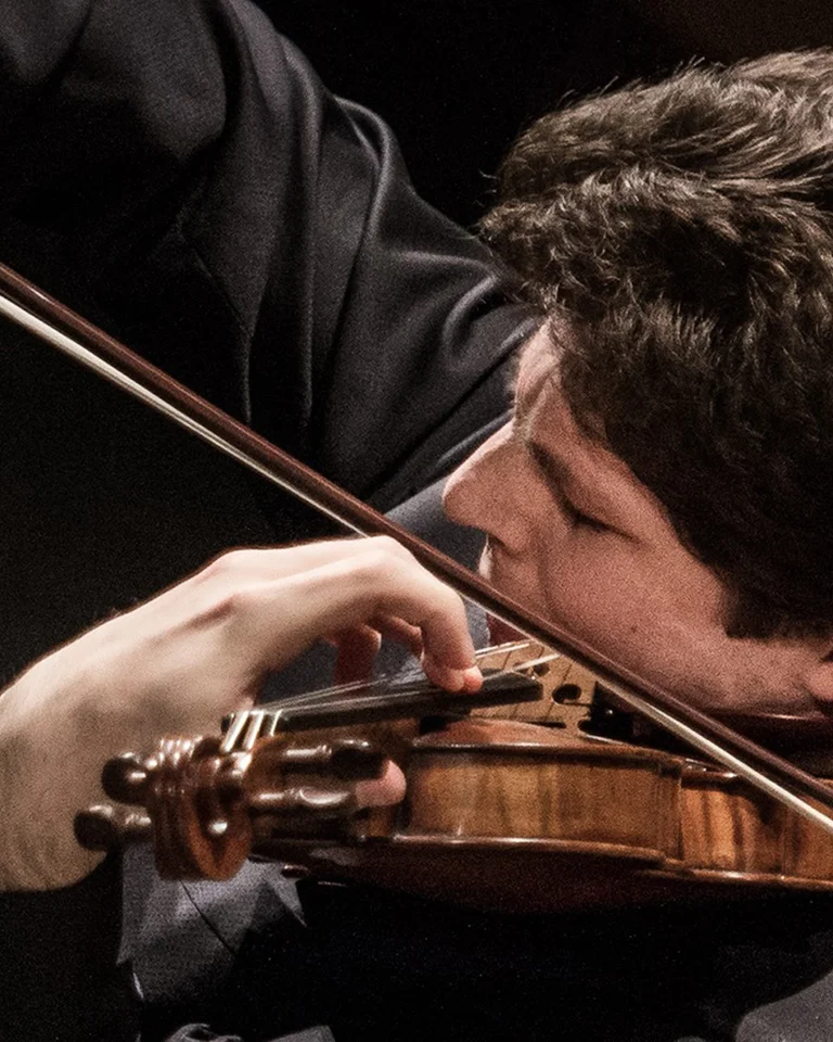 Ein Musiker spielt konzentriert Violine, mit dunkler Kleidung und intensivem Ausdruck, unter gedämpftem Licht auf der Bühne.