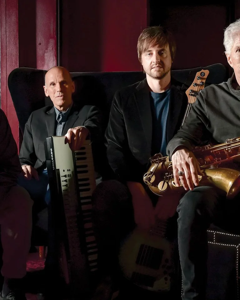 Vier Männer sitzen mit Musikinstrumenten in einem dunklen Raum. Eine entspannte Atmosphäre, die auf eine Jazz-Band hindeutet.