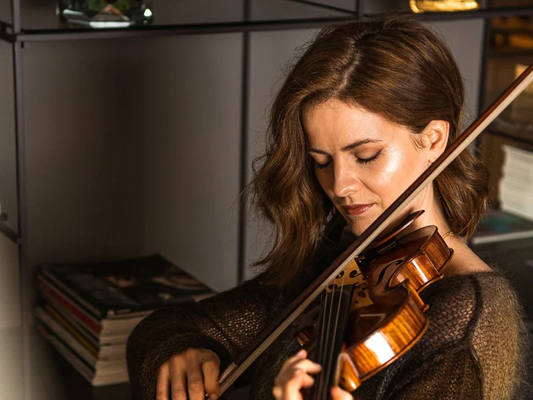 Eine Frau (Veronika Eberle) spielt konzentriert Geige in einem modernen Innenraum. Im Hintergrund sind Bücher und eine Skulptur zu sehen.