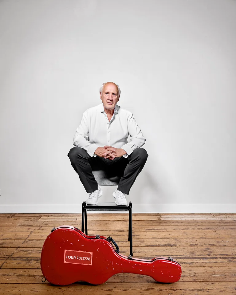 Eine Person (Herman van Veen) sitzt auf einem Hocker in einem leeren Raum. Vor ihr liegt ein rotes Gitarrenkoffer auf Holzboden.