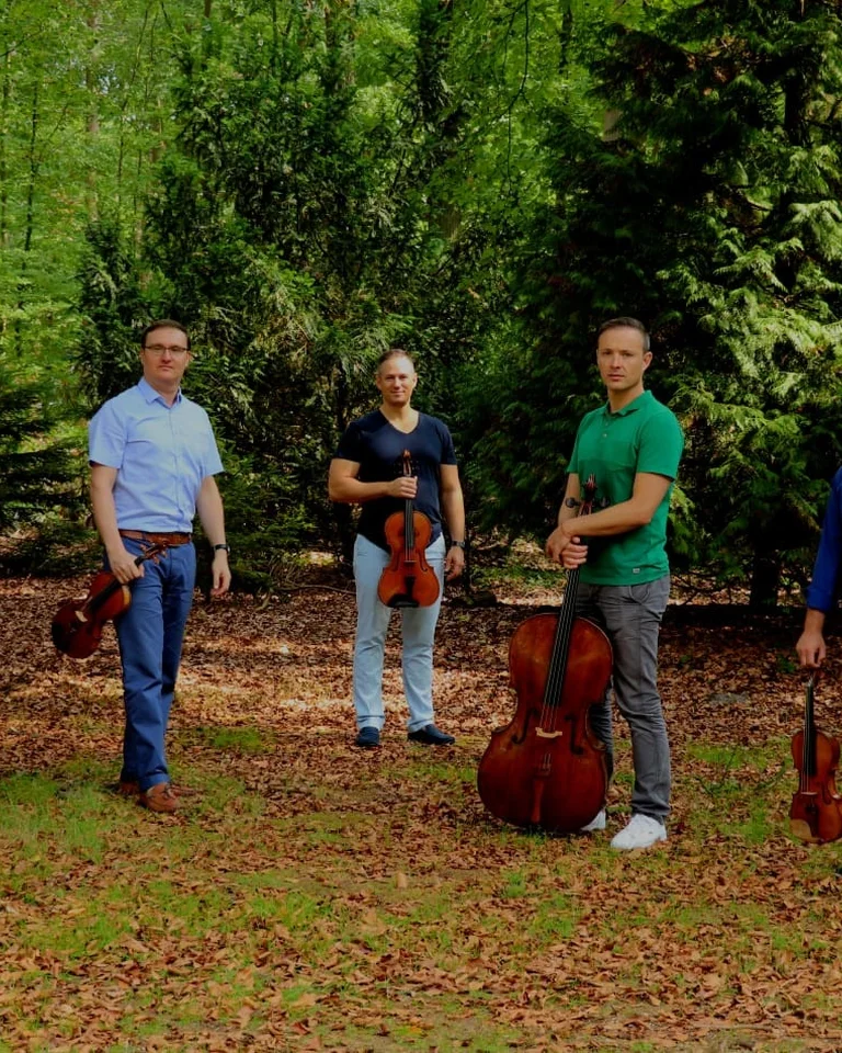 Fünf Musiker stehen mit ihren Instrumenten im Freien. Sie sind von Bäumen umgeben und scheinen sich auf eine Aufführung vorzubereiten.