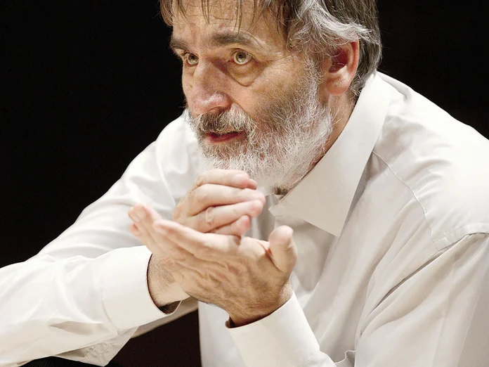Ein Mann mit grauem Bart und weißem Hemd macht eine nachdenkliche Geste mit den Händen. Er wirkt konzentriert.