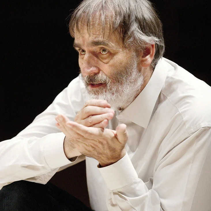 Ein Mann mit grauem Bart und weißem Hemd macht eine nachdenkliche Geste mit den Händen. Er wirkt konzentriert.