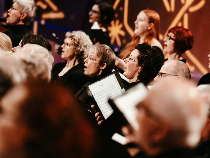 Ein gemischter Chor (Rodenkirchener KammerChor) singt auf einer Bühne. Die Chormitglieder halten Notenblätter und tragen schwarze Kleidung.
