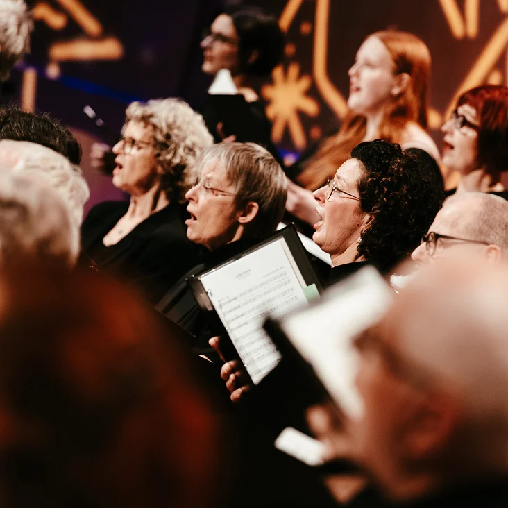 Ein gemischter Chor (Rodenkirchener KammerChor) singt auf einer Bühne. Die Chormitglieder halten Notenblätter und tragen schwarze Kleidung.