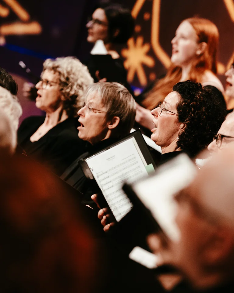 Ein gemischter Chor (Rodenkirchener KammerChor) singt auf einer Bühne. Die Chormitglieder halten Notenblätter und tragen schwarze Kleidung.