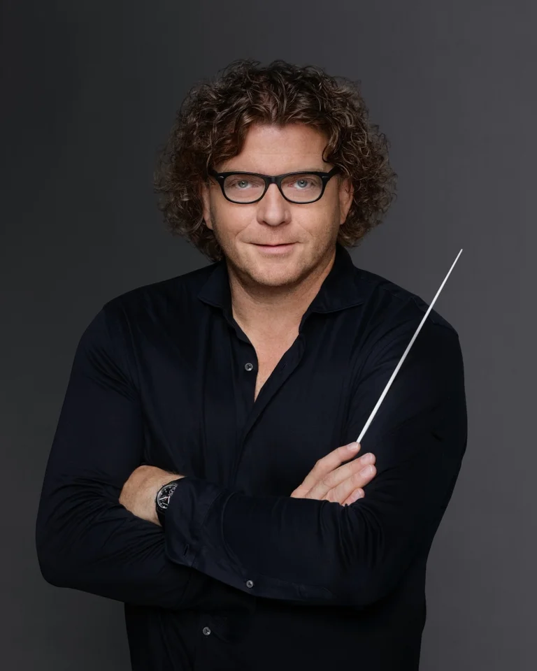 A person with curly hair and glasses holds a conductor's baton, wearing a dark shirt, against a gray background.