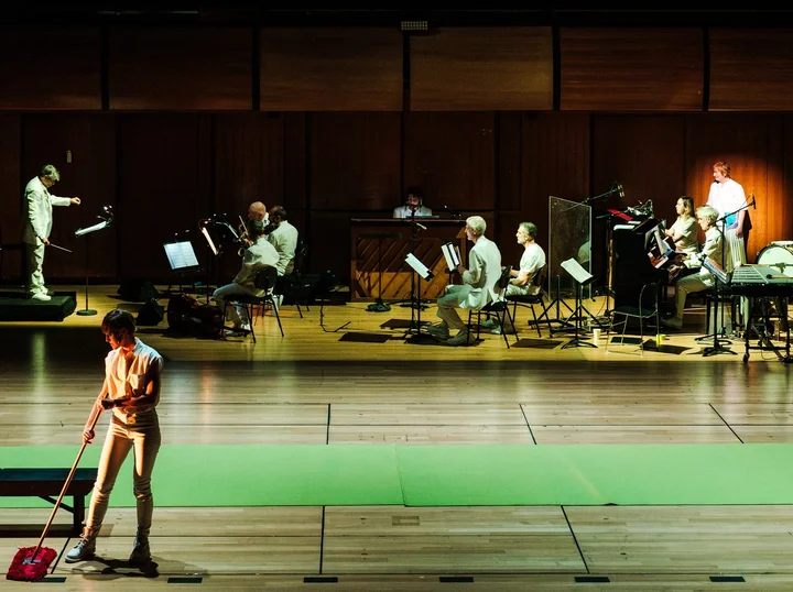 Bühnenszene aus einer Aufführung der "5. Jahreszeit" von novoflot, im Vordergrund ein Schauspieler mit einem Schrubber, im Hintergrund ein Orchester.