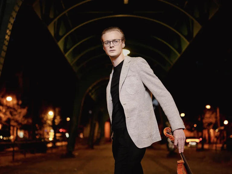 Ein Mann in einem hellen Blazer steht nachts unter einer beleuchteten Brücke und hält eine Geige in der Hand.