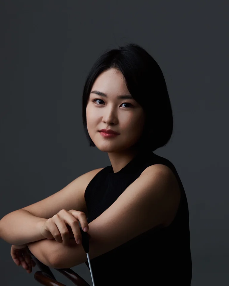 Eine Frau mit schwarzen Haaren und einem schwarzen Oberteil sitzt vor einem dunklen Hintergrund, mit einem sanften Lächeln.