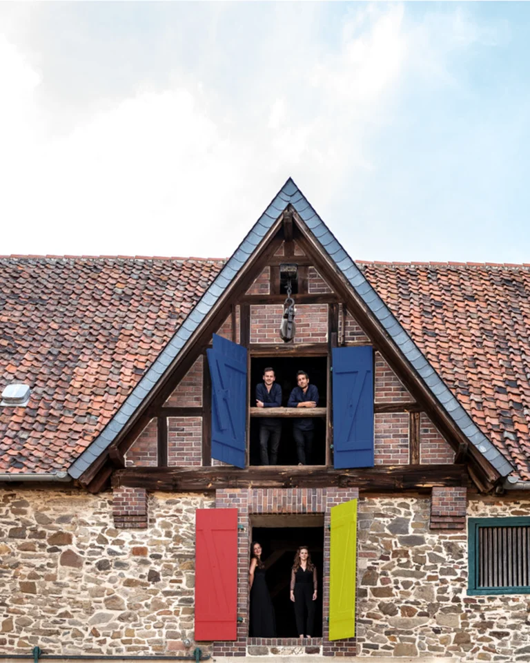 Ein traditionelles Fachwerkhaus mit einem Ziegeldach und farbigen Fensterläden. Menschen stehen in den offenen Fenstern.