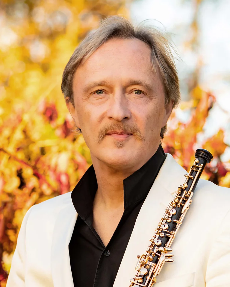 A man in a white suit holds an oboe. The background features autumn-colored shrubs or trees.