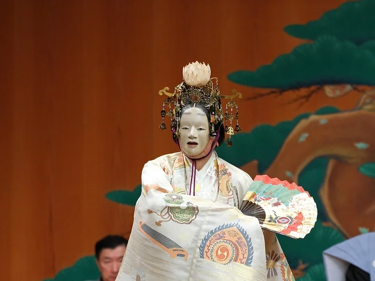 Eine Noh-Darsteller in traditionellem Kostüm (Hagoromo) und Maske führt auf einer Bühne mit Holzhintergrund und gemaltem Baum ein Ritual auf.
