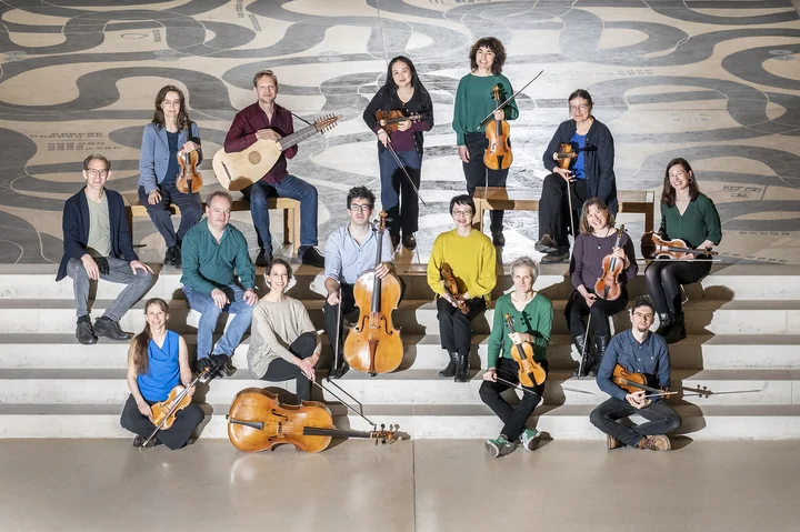 The members of the B’Rock Orchestra are posing on a staircase, some with their instruments. The background features a modern, wavy pattern.