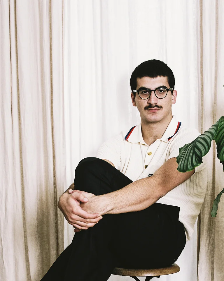 Giorgi Gigashvili sitzt auf einem Hocker in einem Zimmer mit Vorhängen und einer großen Pflanze neben ihm.