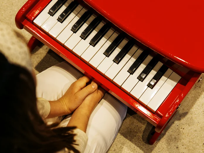 Ein Kind sitzt an einem roten Spielzeugklavier. Die Kamera blickt aus einer Vogelperspektive auf die Tastatur und das Kind.