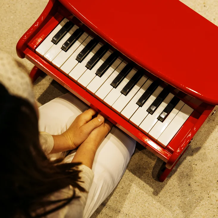 Ein Kind sitzt an einem roten Spielzeugklavier. Die Kamera blickt aus einer Vogelperspektive auf die Tastatur und das Kind.