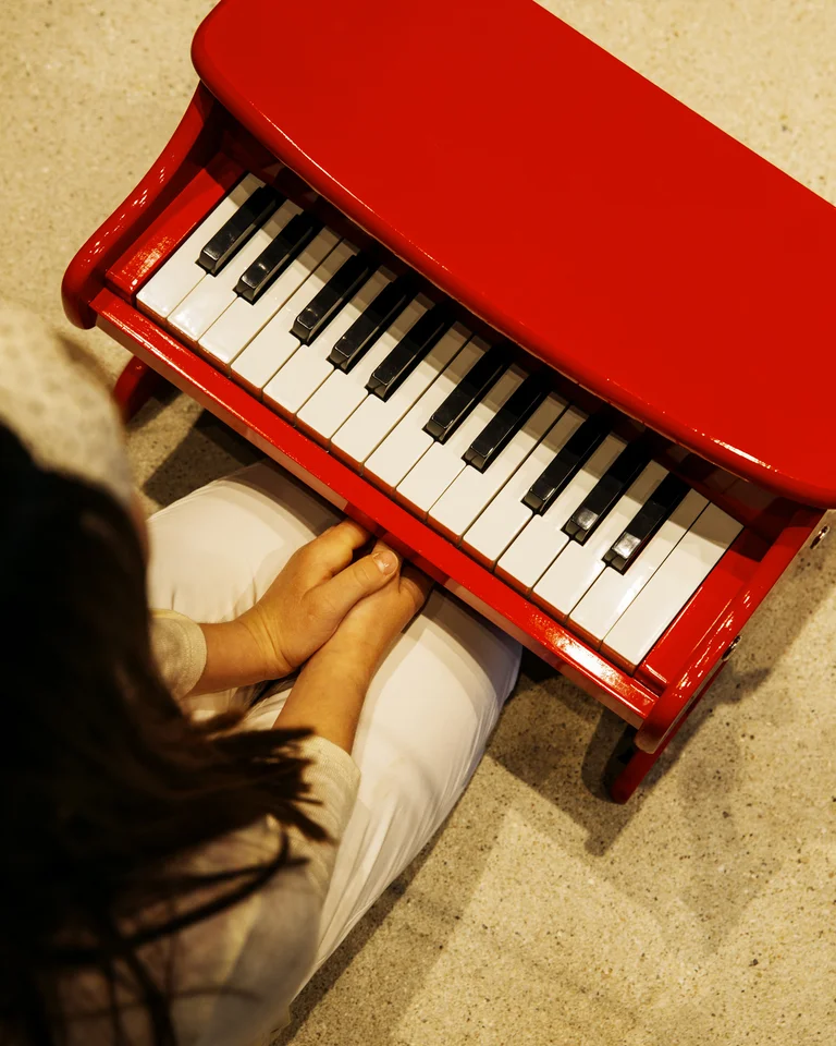 Ein Kind sitzt an einem roten Spielzeugklavier. Die Kamera blickt aus einer Vogelperspektive auf die Tastatur und das Kind.